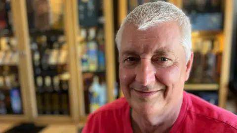 Tom sits in his wheelchair in his living room looking into the camera and smiling. He has short white hair and is wearing a red t-shirt. A wooden display cabinet with a glass front can be seen out of focus behind him. 