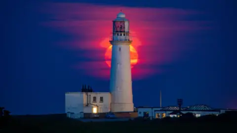 Mark Griffiths A striking red and orange full moon hangs low in the sky behind the stark white of Flamborough lighthouse and associated buildings. 