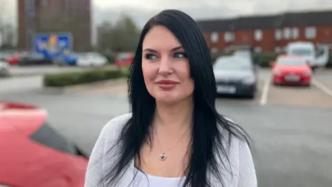 A woman with long dark hair, and a grey top is looking to the left and smiling. She is in a car park, with cars behind her