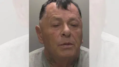 Police custody image of Henderson against a grey background, he has a neck tattoo, dyed black hair and is wearing a grey t shirt