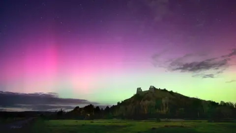 Mathew Browne A large hill is pictured with trees in the countryside, underneath a Northern Lights display in the sky which is illuminating it in pink and green.