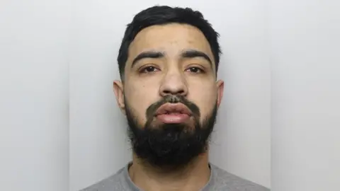 Shazeb Khalid has a blank expression in a police custody photo. He has black hair and a beard.