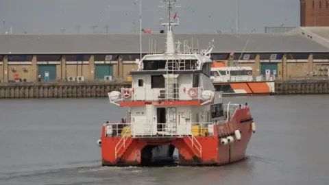 A white boat with a red base is sailing on water in a port, with buildings in the background