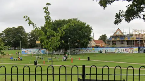 A Google Maps street view image of a metal fence behind which is a recreation ground with a small park. There are swings and a see-saw, as well as large trees and a home that is being constructed.