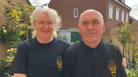 Gerry Orchard and Julie Bushell stand in a garden, with red-brick housing behind them. Gerry has cropped white hair and a black polo with a yellow coffin-shaped Lyke Wake Walk logo on his chest. Julie has grey hair in a ponytail and wears a black t-shirt with a yellow coffin-shaped Lyke Wake Walk logo on the chest.