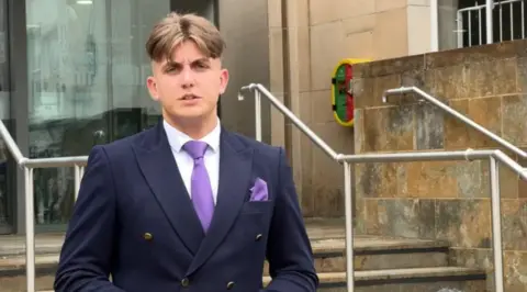 BBC A man with short hair and a navy jacket with a purple tie and handkerchief on the steps of a stone building