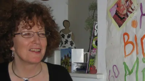 Mick Murray Ann has curly hair and glasses, is smiling, wearing a black top and silver dangling earrings 