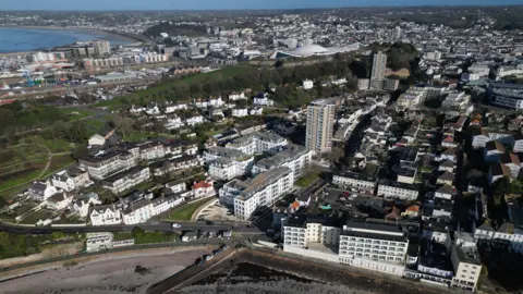 BBC An aerial photo of St Helier. There are a number of large five storey buildings and a few high rise towers. There is also a park visible. There is a bay in the foreground and another in the distance to the left.