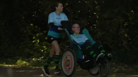 Alice pushes David through the park at speed. David is sat in a special wheelchair. They both wear running clothes including green and black stropy socks and pale blue t-shorts with their names on. David wears glasses, a green neck scarf and has a small grey beard. Alice has brown hair tied back. 