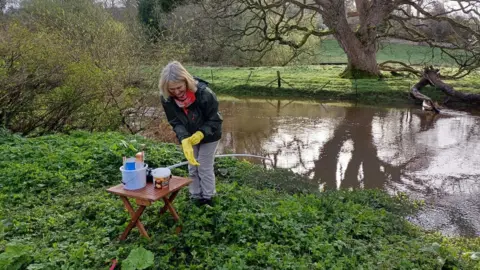 Jane Davis A woman is standing in front of a small foldup table on a riverbank. She is wearing yellow gloves and is about to do some testing 