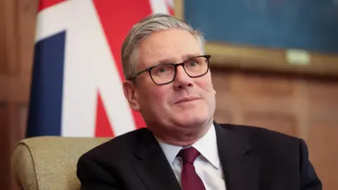 EPA British Prime Minister Keir Starmer is seen sitting in front of the a large Union Jack, wearing a dark jacket, red tie and his customary black rimmed glasses.