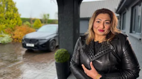 A woman in leather jacket sits in a wheelchair looking into the camera on her front porch. In the background a black jeep is parked in the driveway. 