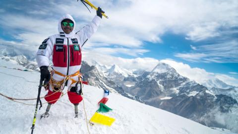 A man with two prosthetic legs wearing red and white snow clothes at the top of Mount Everest