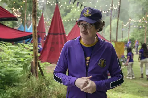 Netflix Actor Steve Buscemi as Principal Barry Dort, he wars a purple sports outfit and cap with yellow badge and trim and dark rimmed glasses.
