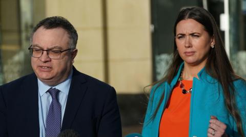 Stephen Farry (left) and Sorcha Eastwood (right) standing in front of media microphones