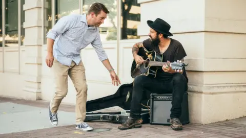 A stock image of a man walking past a busker and dropping money into his guitar case, which is laying on the floor. The man walking by has short light brown hair styled in a side parting. He is wearing a blue and white fine line gingham check print shirt with the sleeves rolled up, a pair of beige chino trousers and blue and white Converse trainers. The busker is a man wearing a wide brimmed black hat, he has a black beard, black short sleeved v-neck t shirts, indigo jeans and weathered brown leather boots. He has tattoos on his arms and is holding a black acoustic guitar on his right knee, sitting on an amp.