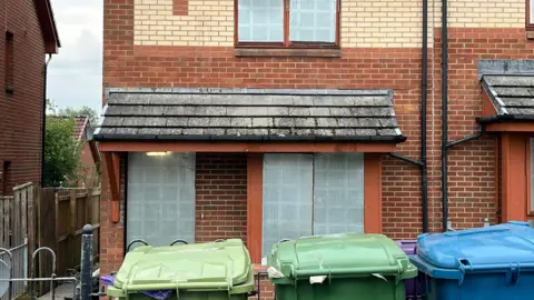 Front view of a two-storey brick house with a tiled roof and a small covered porch. Three wheelie bins, two green and one blue, are positioned in front of the house. The porch roof shows visible moss and weathering, and the windows have closed blinds. A wooden fence and neighbouring houses are visible on either side under an overcast sky.