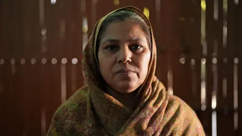 BBC / Neha Sharma Sudesh Kumari stood in her home in Delhi, looking directly at the camera with a neutral expression. She is wearing a patterned headdress