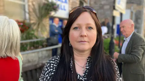 A woman stands outside a town hall style building where people are gathering for a meeting. She is wearing a patterned black and white dress and has sunglasses sitting on top of her head. 