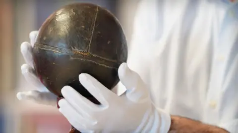 A person wearing white protective gloves holdings a small, shiny leather ball.