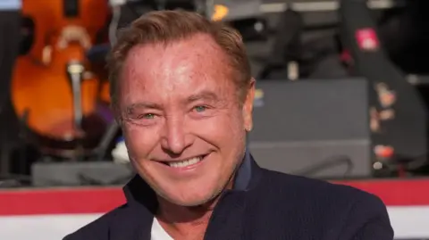Brian Lawless/PA Wire Michael Flatley, a 67-year-old man with short, blonde hair and blue eyes, smiles at the camera. He is wearing a white t-shirt under a navy jacket. The collar of his jacket is turned up. There is a stage behind him on which sit a double bass, instrument cases and large black speakers.