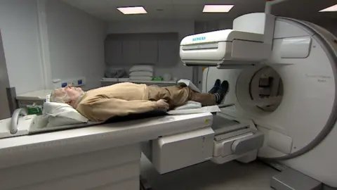 A man lies on a medical scanner bed, in hospital. He is wearing brown trousers and a light brown jumper, and shoes and socks. His feet are about to enter a small round opening in a large white device with an flat camera overhead.