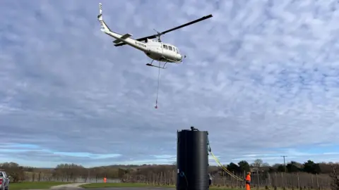 Chrissie Reidy/BBC A helicopter picking up the treatment tank to be taken to the Southern Water wastewater treatment works in Pembury. Each tank had to first be picked up from an adjacent orchard, moving to hover above Pembury Wastewater Treatment Works