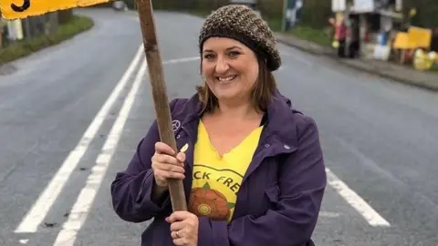 Claire Stephenson Claire Stephenson standing in a street holding the handle of a placard. She has brown, straight, shoulder-length hair under a brown woolly hat and is wearing a yellow "frack free" t-shirt with a red rose and purple coat.