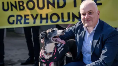 Mark Ruskell, who has short grey hair, kneels beside a black and white greyhound turned towards him with its mouth wide open. Ruskell, wearing a suit, his smiling at the camera. A yellow protest banner is held in the background, 
