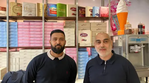 Steve Jones/BBC Barber Rashid stood with a member of staff at his restaurant, with rows of confectionery and an ice cream machine behind them