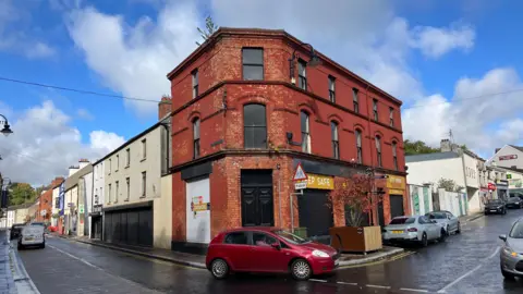 The town centre of Dromore in County Down.  There is a red brick building on a corner site in the foreground - it has a black door and looks boarded up. There is a red car in front of it turning the corner. The building next to it is white and the black shutters are down