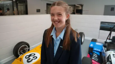 Student Clara smiling for the camera. There are racing cars behind her.