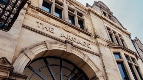 A stone Victorian archway with decorative iron bars inside. Above the archway is The Arcade engraved into the stone masonry. There are windows in the building with stone mullions.