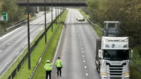 A white, Scania lorry, with a forklift on the back, is stopped on the A63. Behind it are tyre marks and debris. A bridge can be seen in the background. Two police officers are looking at the scene.