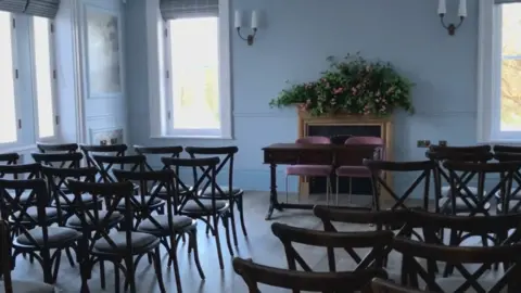 A light blue painted room with large windows, and lots of wooden seats facing a top table with two seats.