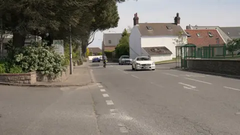 A road in St Peter, with a petrol station on the left hand side of the road - largely covered in trees, and houses and a bus station on the right hand side. There is a right-hand turn lane in the middle of the road. A white car is indicating into the turn lane, with a silver car travelling behind it. A cyclist and blue car are on the left hand side of the road travelling away from the camera.