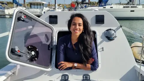 Ananya is smiling as she leans out the enclosed, 'indoor' space of her boat. It has a square, hatch-like window.