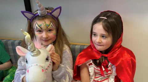 Two young girls sitting down wearing World Book Day costumes.  Rachel has long blonde hair and is wearing a purple unicorn headband, a light purple cardigan and she is holding a large white unicorn toy.  Ava has long dark hair and is wearing a red cape and a red and and white dress. 