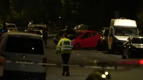 Cameron Davidson/BBC Police officers at the scene of a crash at night. A red car's bonnet is damaged.