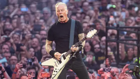 A white haired man wearing all black and pulling a screaming face as he plays electric guitar on a stage, surrounded by huge crowds, many of whom are holding up their arms or phones.