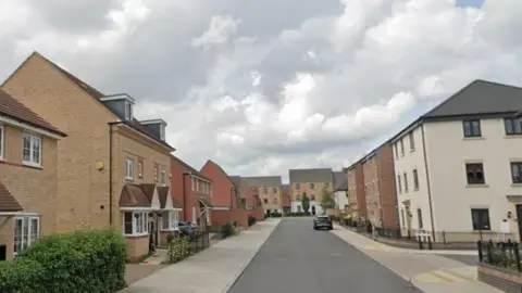 Google A new housing estate showing two and three-storey brick-built houses around a wide grey road with cream-coloured pavements. A car is parked outside one house. There are dark-coloured railings outside some houses and hedges in some places.