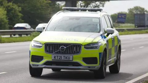 A police car is parked on a three-carriageway road. It is yellow and blue.