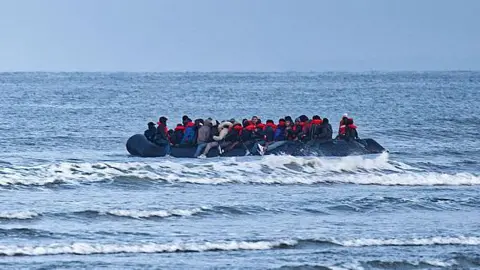 An overloaded inflatable boat sails through waves on a grey day. 