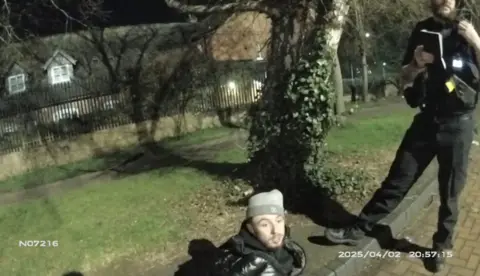 Nottinghamshire Police Body cam image of Bullivant, sat on the ground in a park, with an police officer stood next to him using his radio