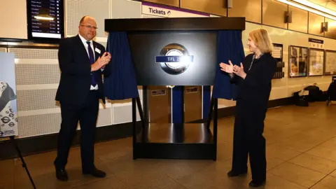 TfL London's Transport Commissioner Andy Lord (left), a man with glasses wearing a black suit, white shirt and blue tie and London Transport Museum CEO Elizabeth McKay (right), a woman with short light hair wearing a black outfit, unveil a silver TfL 25 Roundel inside a TfL station