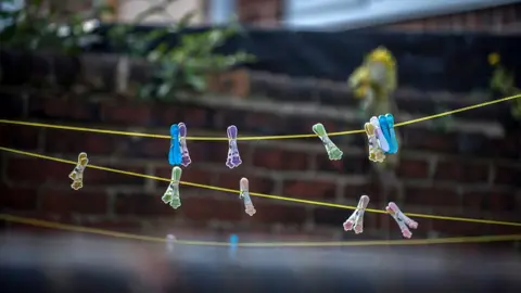 Three criss-crossing strings of yellow clothes line with clothes pegs in assorted pastel colours - blue, pink, purple and green. Beyond is the red-brick wall of a back yard, slightly out of focus.