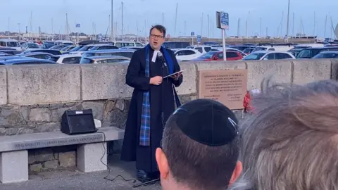 Reverand Justin Taylor leading the Memorial Service, stood next to a plaque to the Guernsey Eight. In the foreground there is an attendee wearing a traditional Jewish kippah.