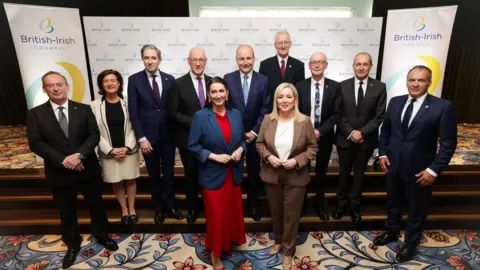 Political leaders from across Ireland, the UK and Crown Dependencies post for a photograph on a set of carpeted steps inside a building during the British-Irish Council summit. Hoardings are behind them with the British-Irish Council logo on.