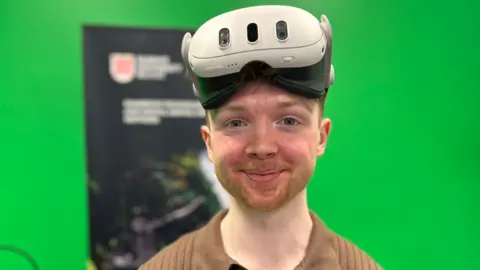 A man with red facial hair and wearing a white VR headset smiles at the camera. He is wearing a brown shirt. He is standing in front of a Queen's University Belfast banner and a green screen.