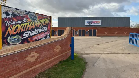 British Speedway External view of Northampton Shaleway, showing the back of a stand, car park and signage.